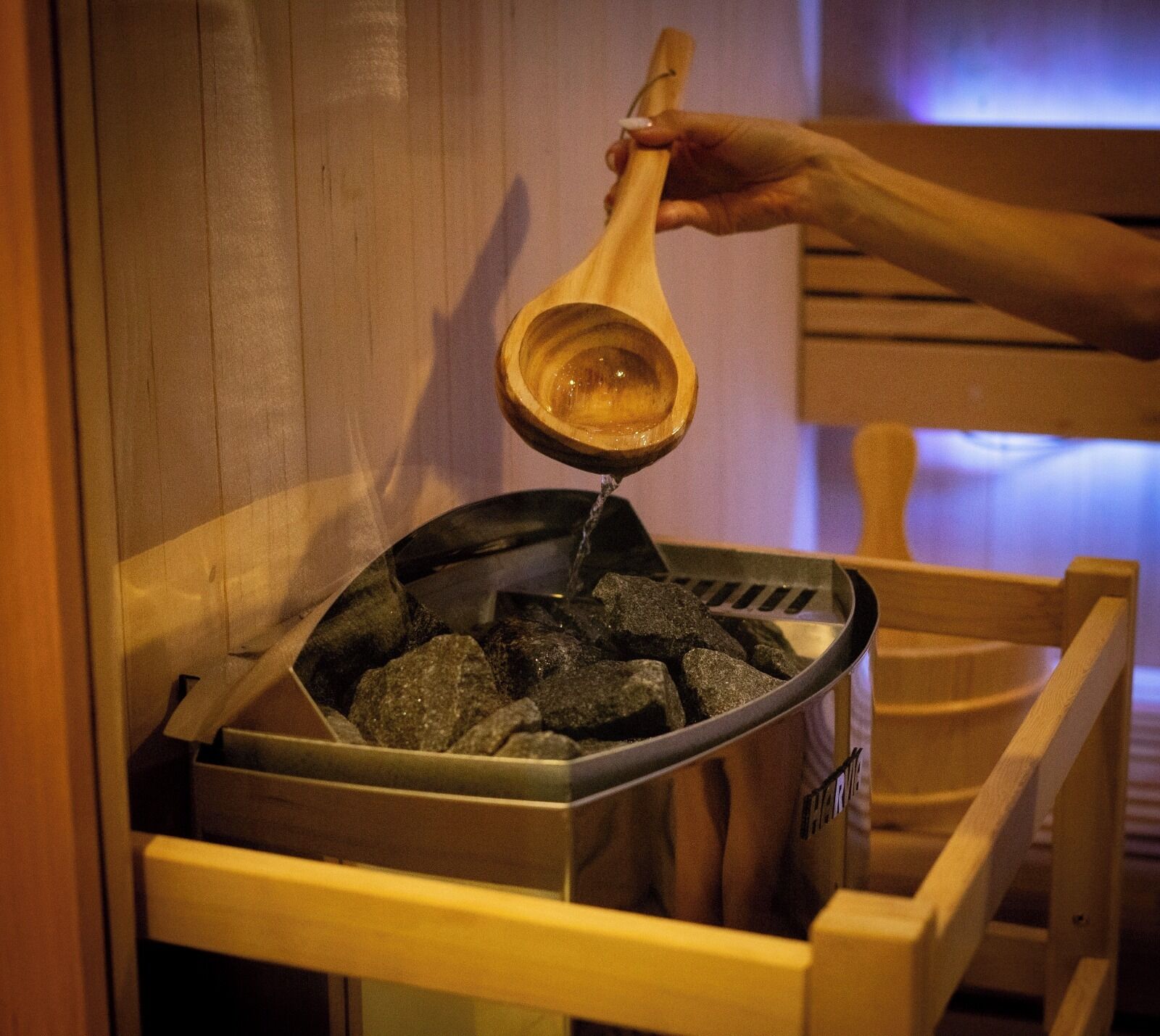 A person pouring a liquid from a wooden ladle into a metal container filled with rocks.
