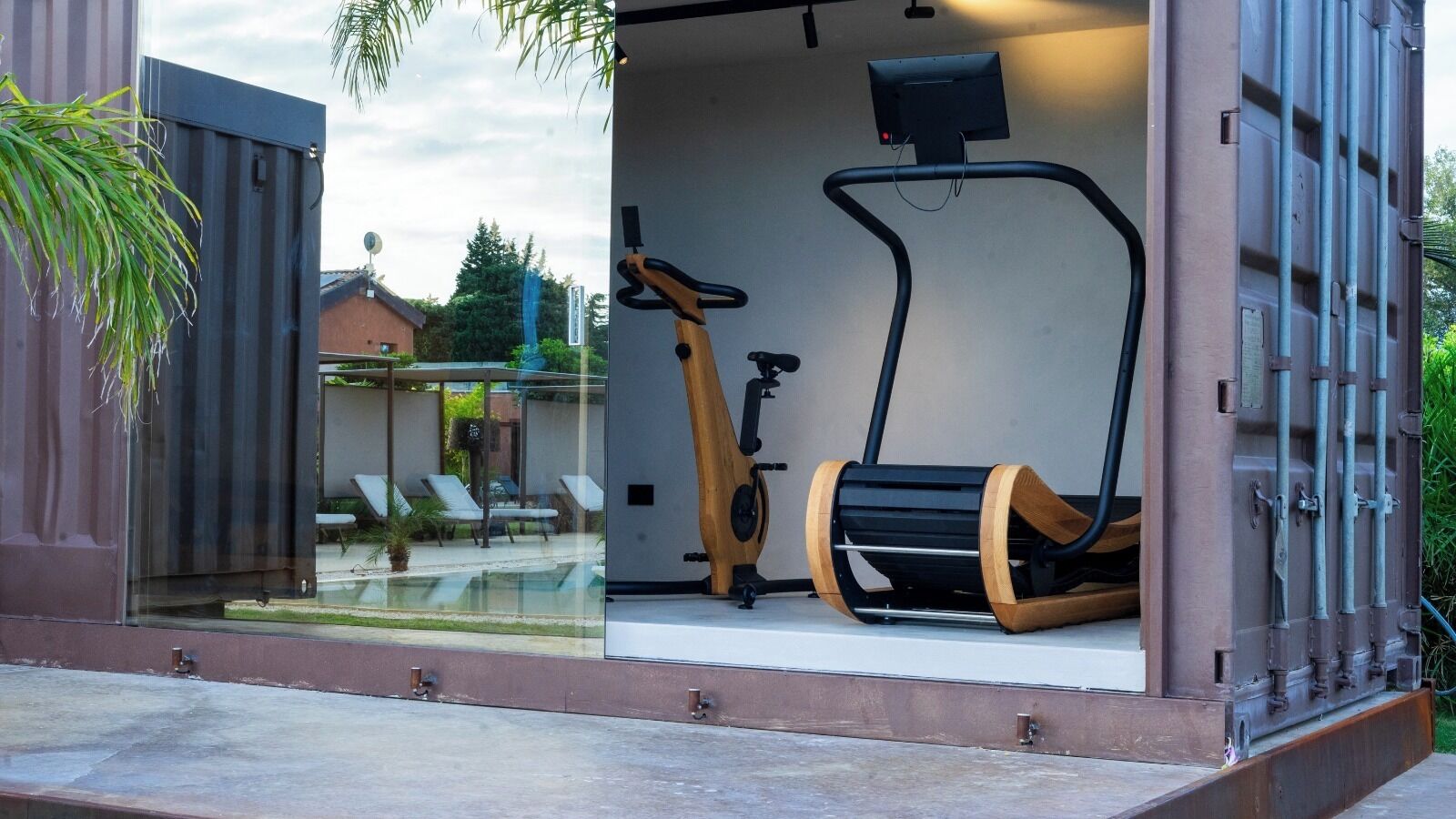 A modern outdoor gym with exercise equipment, including a stationary bike and elliptical, enclosed by glass walls.