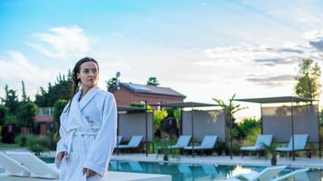 A person in a white bathrobe standing by a pool with lounge chairs and a covered area in the background.
