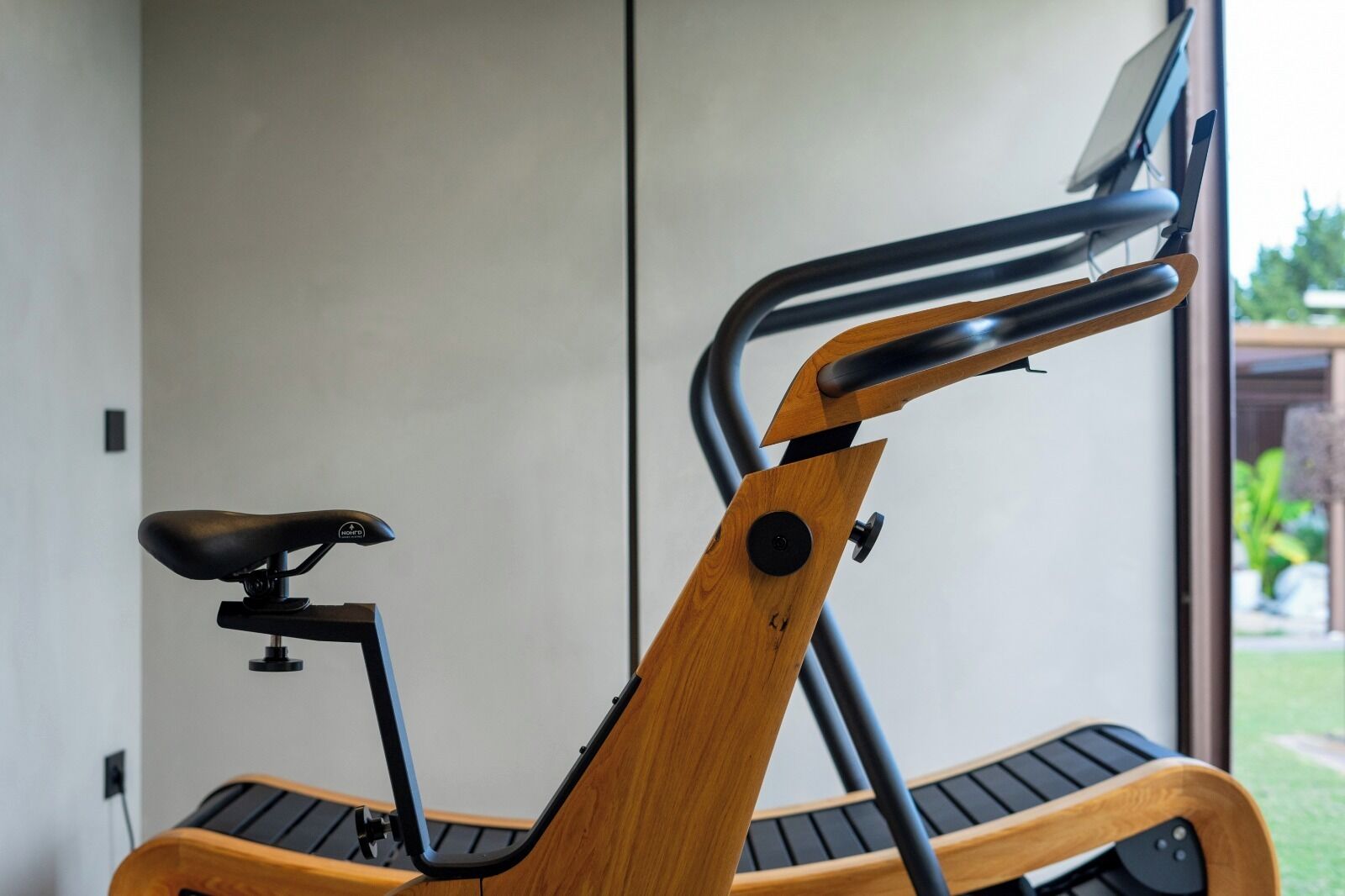 Ein stehendes Ergometer mit Holzrahmen und schwarzem Sattel, platziert in einem Innenraum vor einer schlichten Wand und einem Fenster.