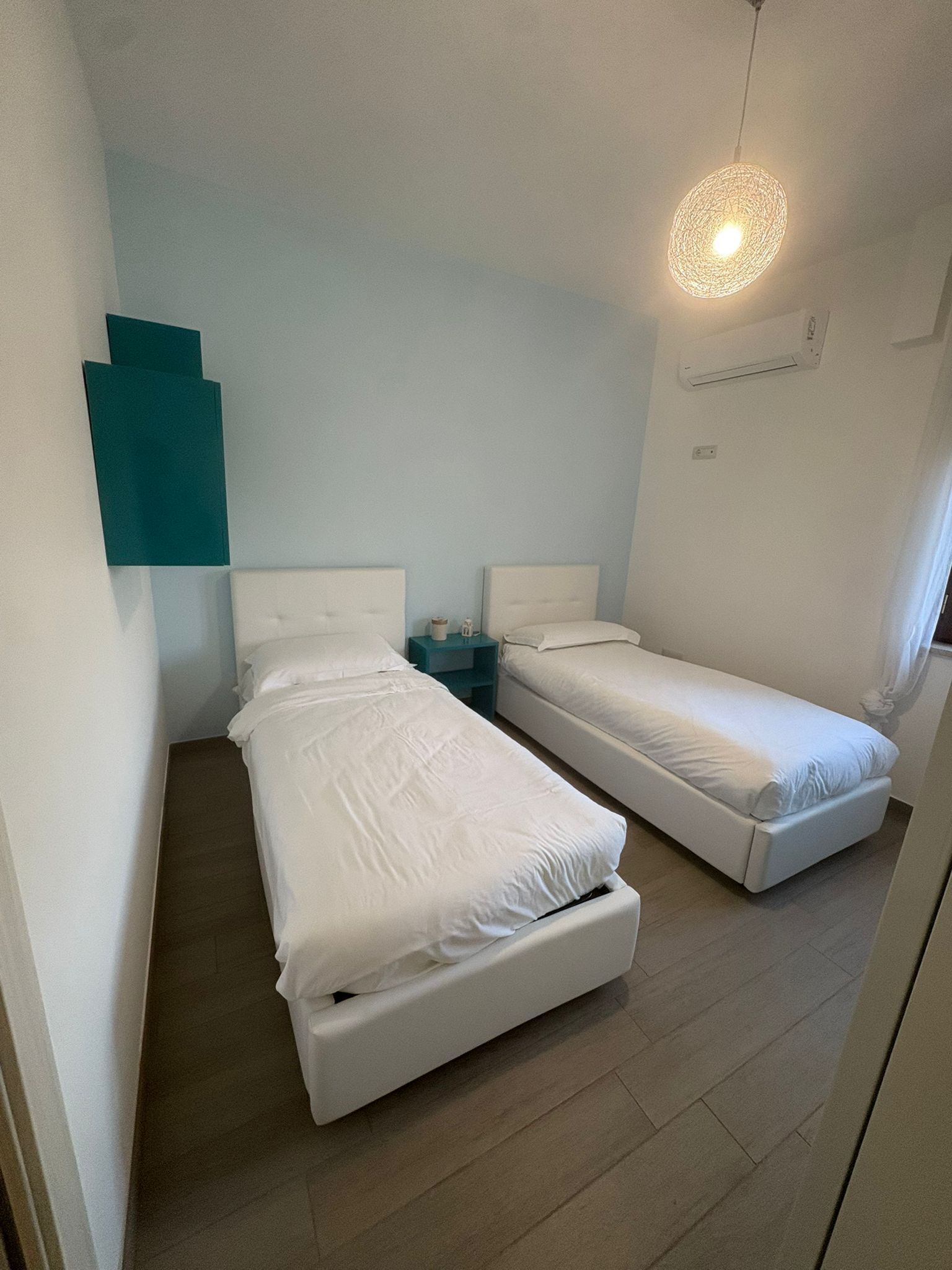 A room with two beds, a teal nightstand, a wall-mounted light fixture, and a wooden floor.