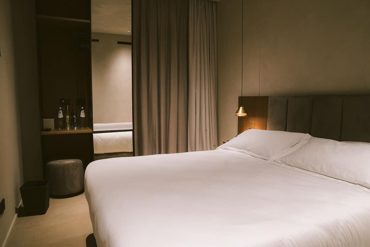 Een hotelkamer met een groot bed, een nachtkastje met flessen, een klein krukje en een gordijn.