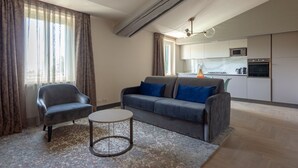 A modern living room with a grey sofa, a small round table, and a chair. A kitchen with white cabinets and appliances is visible in the background. - Residence  Tulipe N3004 (Lucca)
