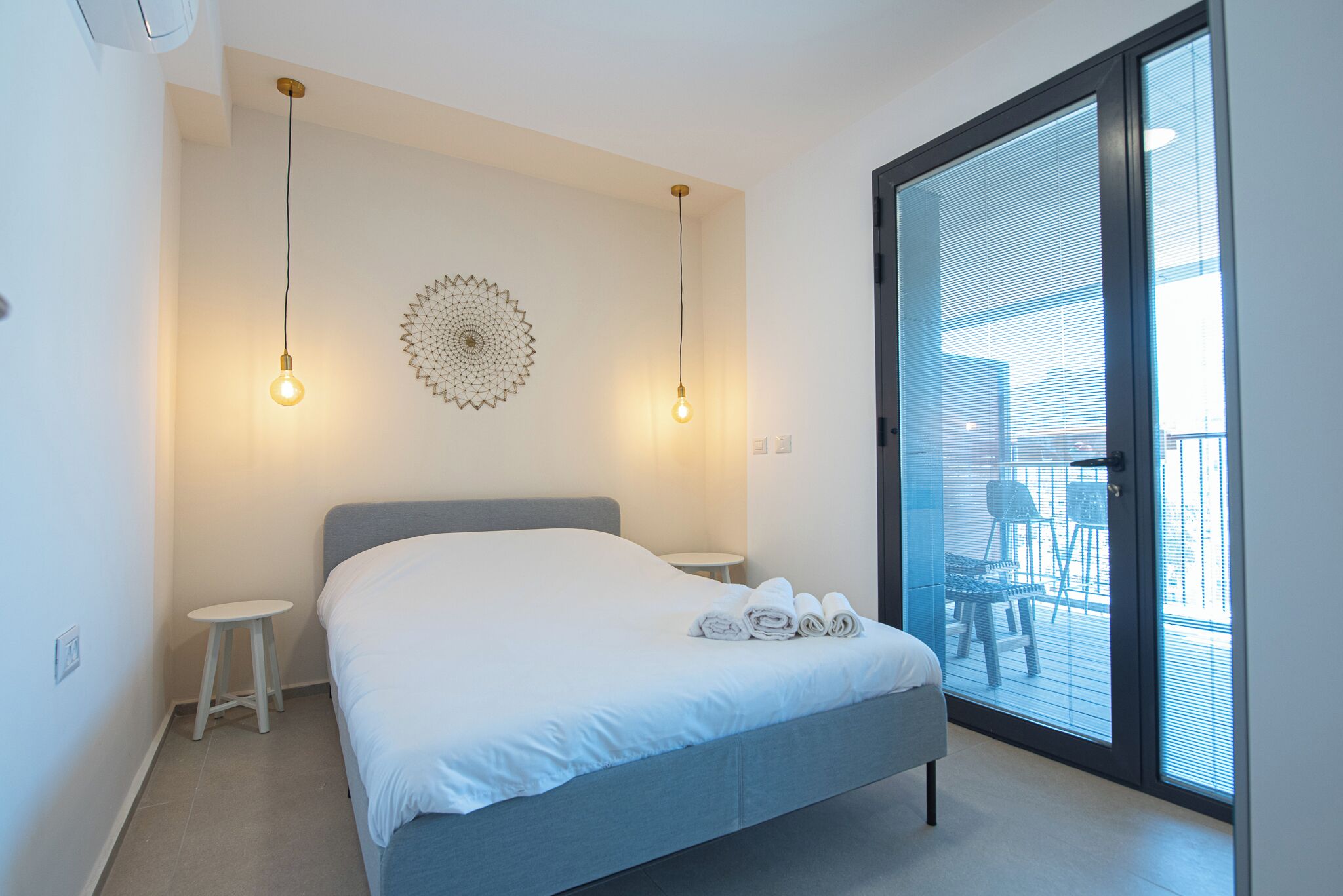 A bedroom with a bed, two bedside tables, a decorative wall piece, and a view through a glass door to an outdoor area with chairs.