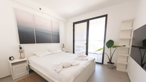 Un dormitorio moderno con una cama grande, una mesita de noche, un televisor montado en la pared, una planta en maceta y un ventanal corredero al balcón.