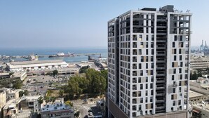 Un edificio de gran altura con numerosos pisos, ventanas amplias y vistas a un puerto con barcos y grúas.