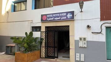 L’entrée d’un hôtel, avec une enseigne en arabe et en anglais, une porte grillagée et un bac à fleurs orné de grandes feuilles.