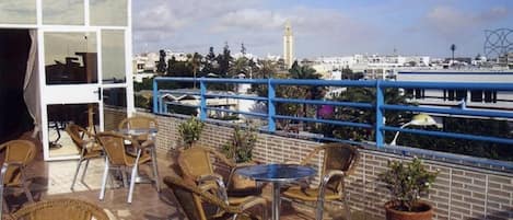 Une terrasse sur le toit, dotée de chaises et de tables en osier, d’une rambarde bleue et offrant une vue sur le paysage urbain, notamment sur une tour.