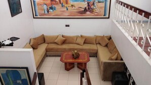 Una sala de estar con un sofá color beige, una mesita de centro con un jarrón, y un cuadro en la pared que representa una escena histórica.