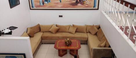 Una sala de estar con un sofá color beige, una mesita de centro con un jarrón, y un cuadro en la pared que representa una escena histórica.