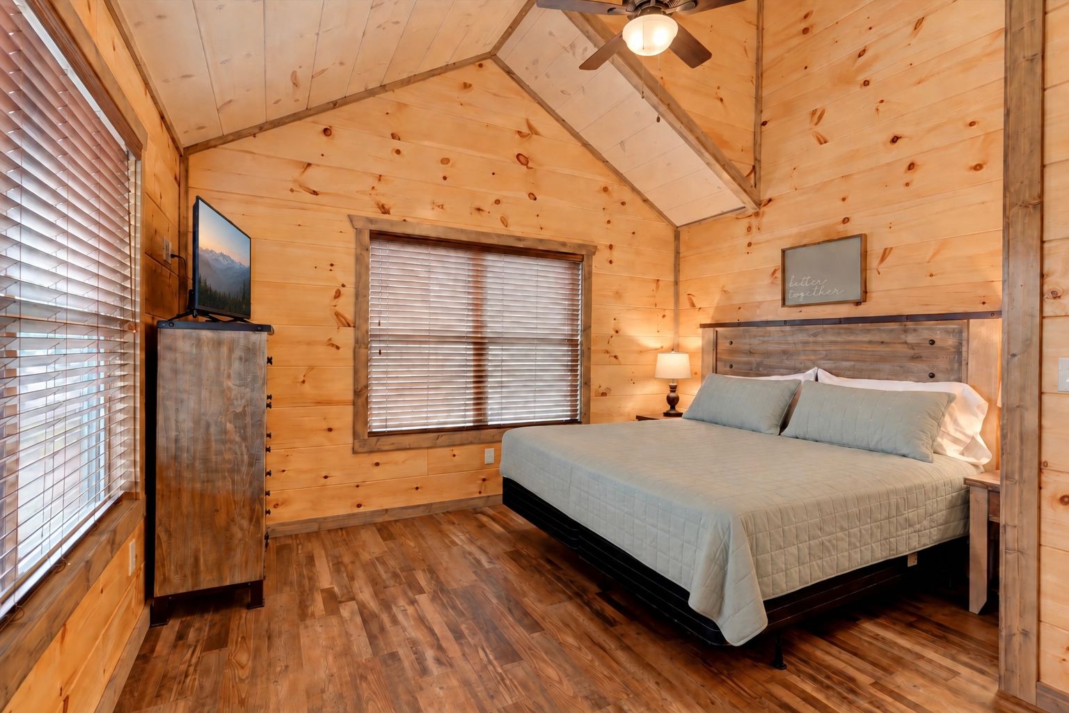 A wooden cabin bedroom with a bed, a window with blinds, a ceiling fan, and a television on a stand.