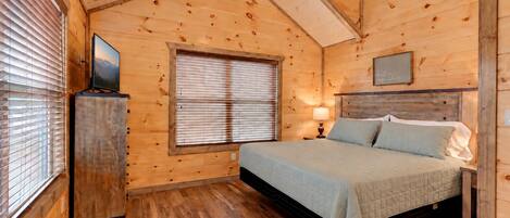 A wooden cabin bedroom with a bed, a window with blinds, a ceiling fan, and a television on a stand.