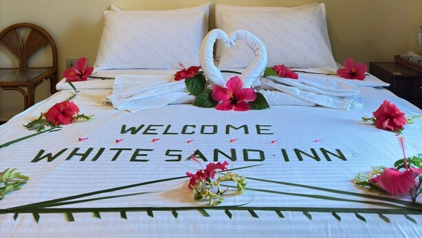 A hotel room with a bed featuring a 'Welcome White Sands Inn' sign, a wall-mounted air conditioner, and a small bedside table.
