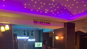 Uma área de recepção com um letreiro 'RECEPTION', um balcão de recepção e um letreiro 'Scammar Hotel' ao fundo.