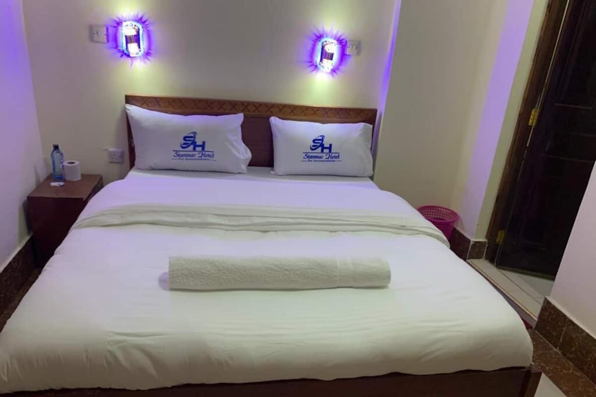 A neatly made bed with white linens and a rolled towel, two pillows with the hotel logo, and two wall-mounted lights.