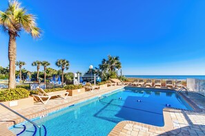 A clear blue pool with lounge chairs and palm trees, surrounded by a fence and a view of the ocean.