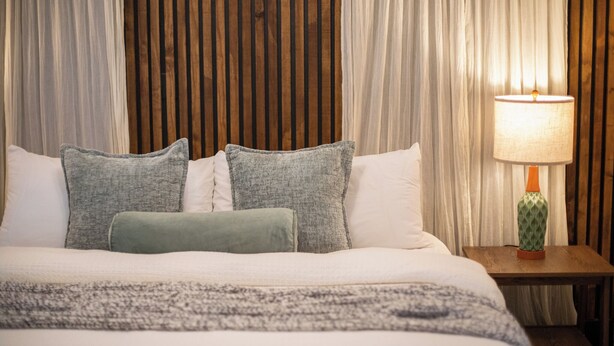 Ein ordentlich bezogenes Bett mit Kissen, eine Lampe auf einem Nachttisch und ein Holz-Kopfteil.