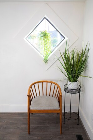 Una silla de madera con un cojín, una planta en maceta sobre un soporte de metal negro, y una ventana con una planta colgante.