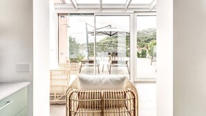 Ein heller Raum mit einer großen Glasdoor, die auf eine Terrasse im Freien führt. Im Raum steht ein Korbstuhl und ein kleiner Tisch.