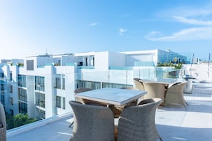 A rooftop terrace with glass railings, wicker furniture, and a clear view of the cityscape.