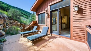 A wooden cabin with a patio, two lounge chairs, a stone wall, and a view of a forested hillside. - 2430 Deer Valley Getaway! Peaceful Home Minutes From Deer Valley With Private Hot Tub! 4 Bedroom Home by RedAwning (Park City)