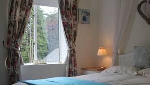 A bedroom with a bed, a nightstand with a lamp, a window with floral curtains, and a painting on the wall.