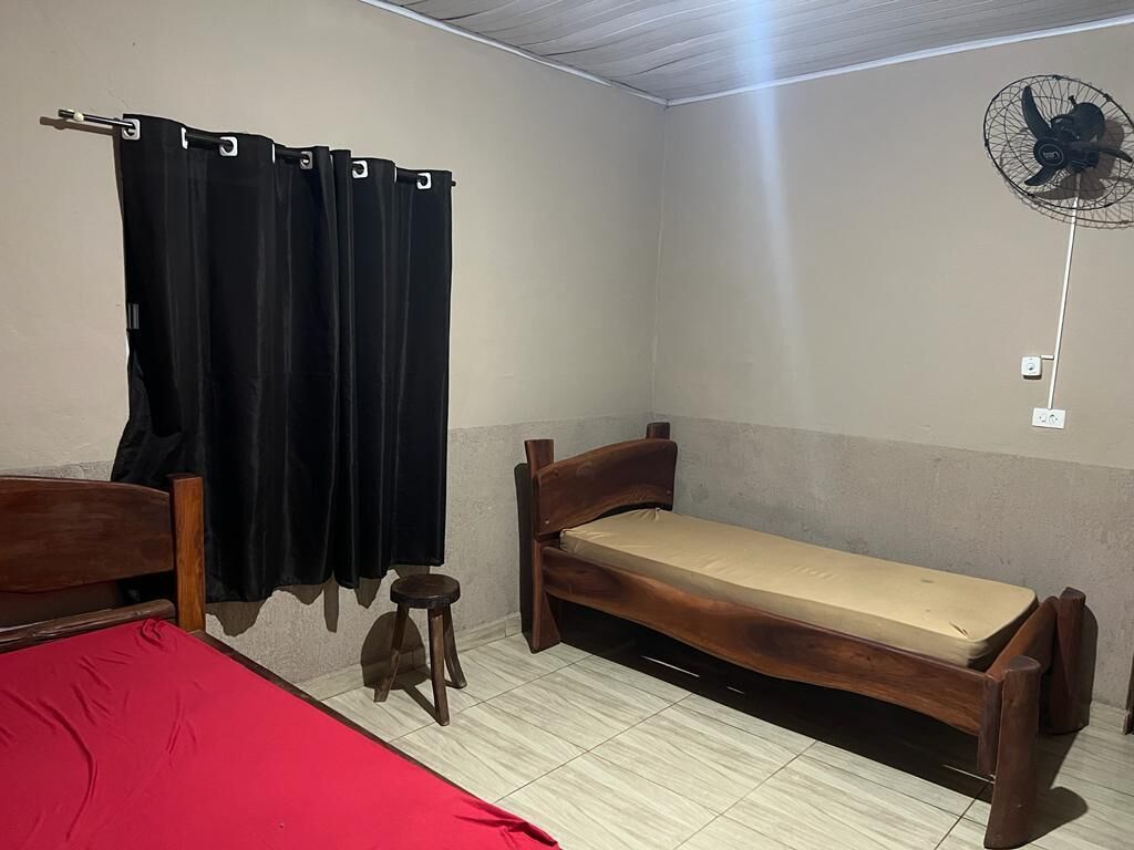 Una stanza modesta con un letto di legno, un materasso rosso, una panca di legno, uno sgabello e una tenda nera, oltre a una ventola a soffitto.