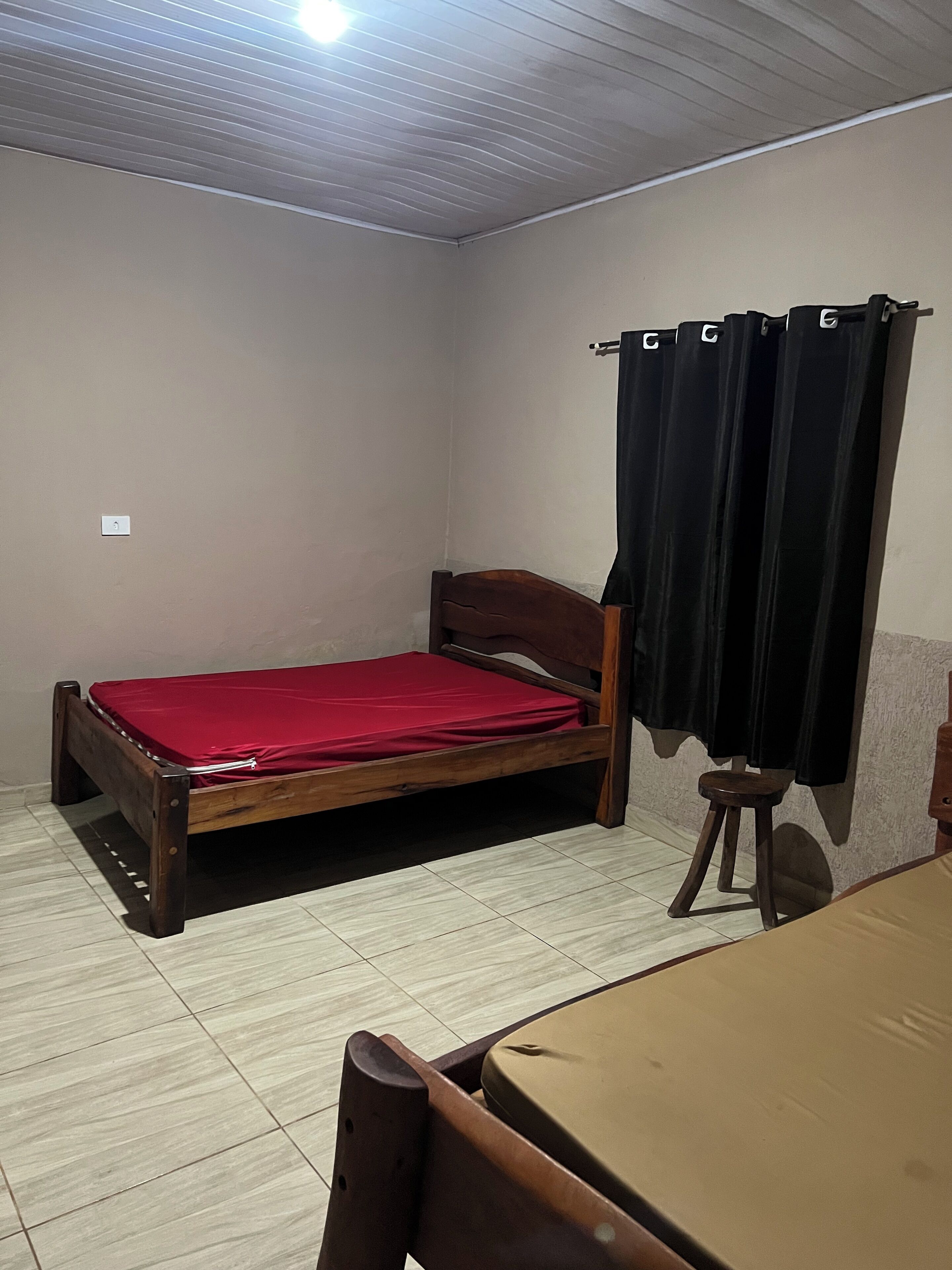A small, single-bed room with a wooden frame, a red mattress, a wooden table, and a black curtain.