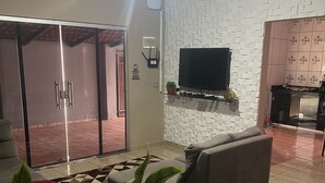 A modern living room with a grey sofa, a flat-screen TV mounted on a textured wall, and a sliding glass door leading to an outdoor area.