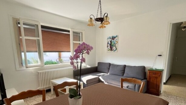 Ein Wohnzimmer mit Esstisch, Stühlen, einer Couch, einem Heizkörper und einem Fenster mit Jalousien.