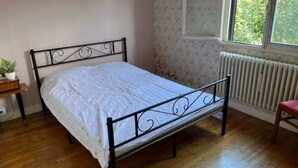 Ein Einzelbett mit Metallgestell in einem Zimmer mit Holzboden, Heizkörper und einem Fenster mit Blick auf Grünfläche.
