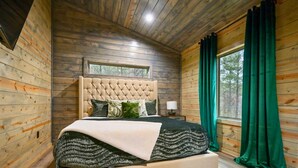 A bedroom with a tufted headboard, a large window with curtains, and a bed with a patterned blanket. - Secluded Forest Retreat with Game Lounge (Broken Bow)