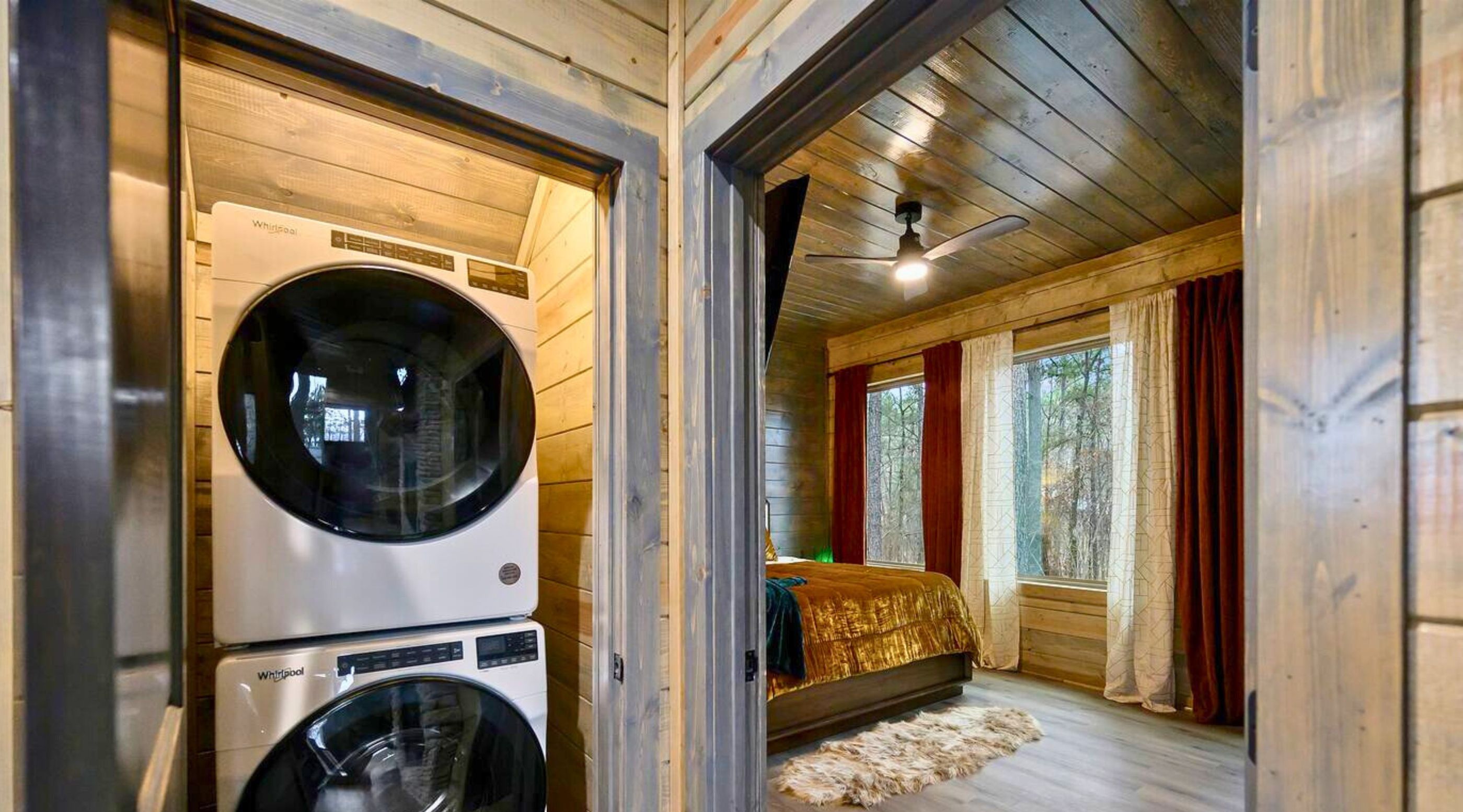 A laundry room with two stacked washing machines and a bed in a room with wooden walls and a ceiling fan.