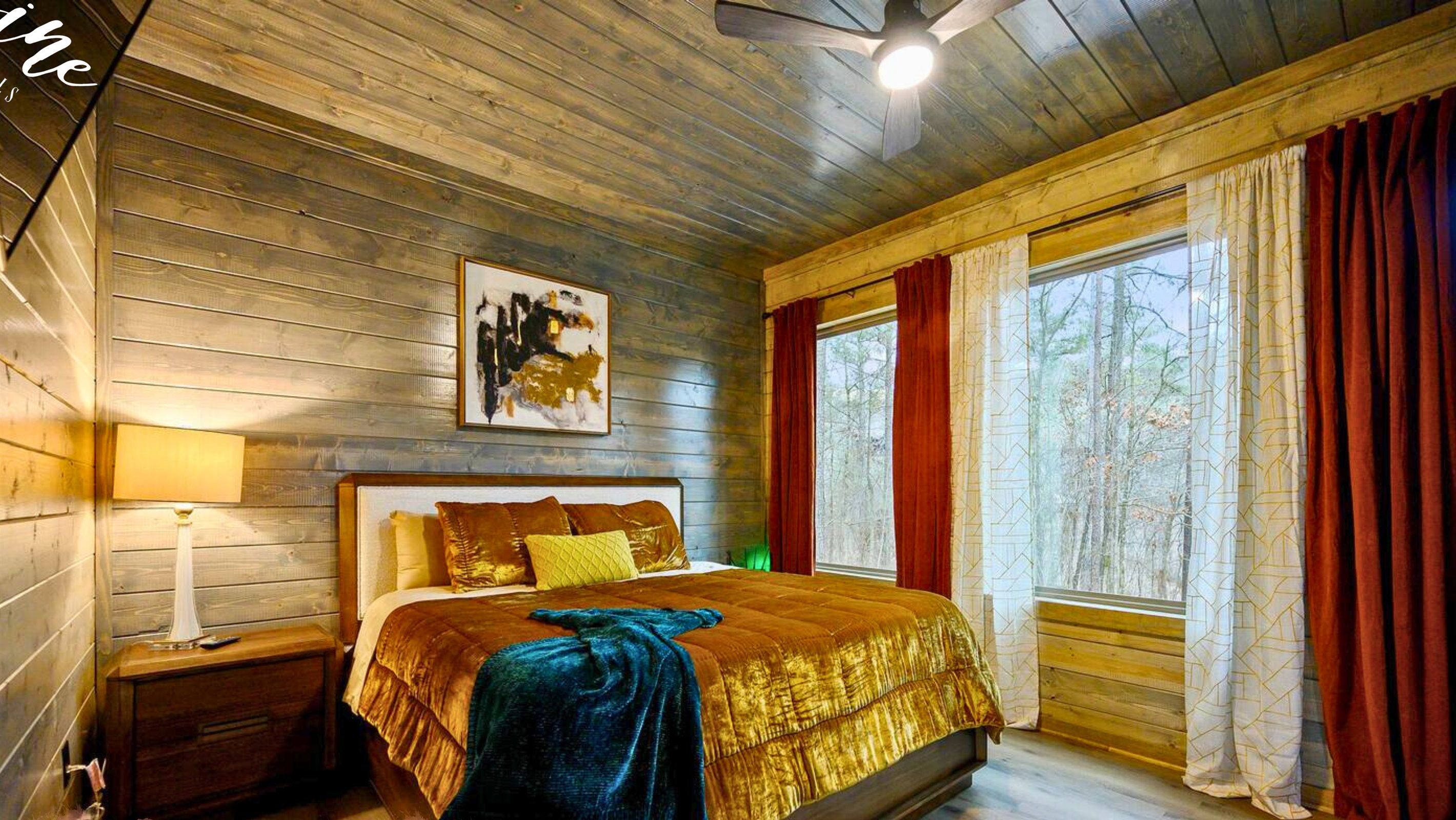 Une chambre à coucher dans une cabane en bois, avec un lit, une table de chevet, une lampe, un tableau et une grande fenêtre avec des rideaux.