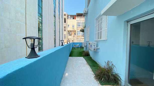 Een smal balkon met een blauwe reling, een klein tafeltje en een stoel. Er staan airconditioners op de muur en een raam met een wit rooster.