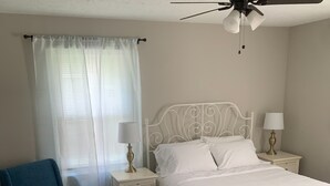 Un dormitorio con una cama de metal blanco, dos mesitas de noche con lámparas, un sillón azul y un ventilador de techo.