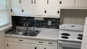 Una cocina con armarios blancos, un lavamanos de acero inoxidable, una estufa y un salpicadero de azulejos.