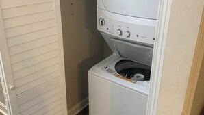 Un cuarto de lavandería con lavadora y secadora de carga frontal, una rejilla de ventilación sobre la secadora y una puerta a la izquierda.