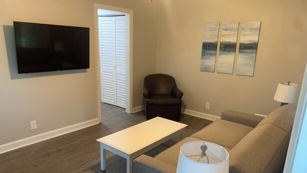 Una sala de estar con televisor de pantalla plana, sofá, mesa de centro y una silla. Se ve una puerta que conduce a otra habitación.