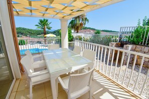 Área externa coberta com mesa de vidro e cadeiras de vime branco, com vista para uma piscina e paisagem deslumbrante.