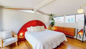 Une chambre à coucher avec un grand lit, une tête de lit rouge, une chaise blanche, une commode en bois et une plante.