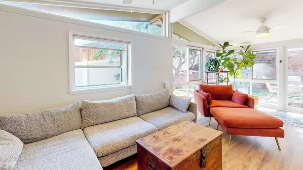 Un salon moderne avec un canapé d’angle, une table basse en bois et un fauteuil rouge. De grandes fenêtres laissent entrer la lumière naturelle et offrent une vue sur l’extérieur.
