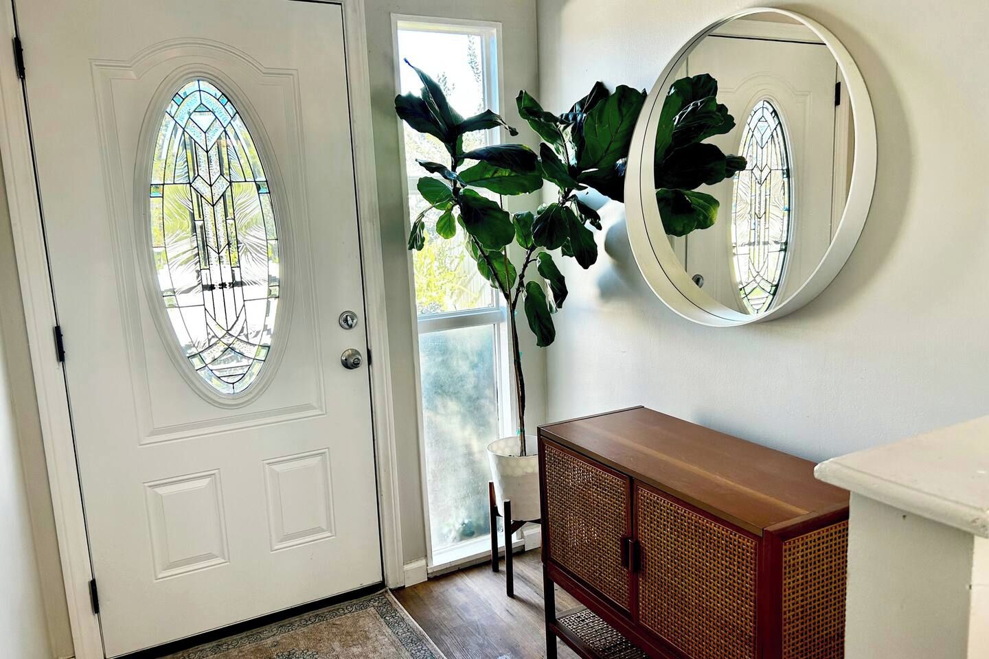 A white door with a stained glass window, a potted plant, a round mirror, and a wooden cabinet.