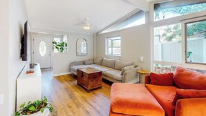 Un salon spacieux doté d’un plancher en bois, d’un canapé d’angle beige, d’un canapé rouge, d’une commode en bois et d’un téléviseur fixé au mur.