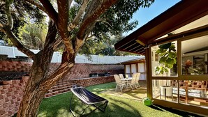 Un jardin avec un hamac, un mur en briques, une terrasse avec des chaises et une porte vitrée donnant sur une pièce.