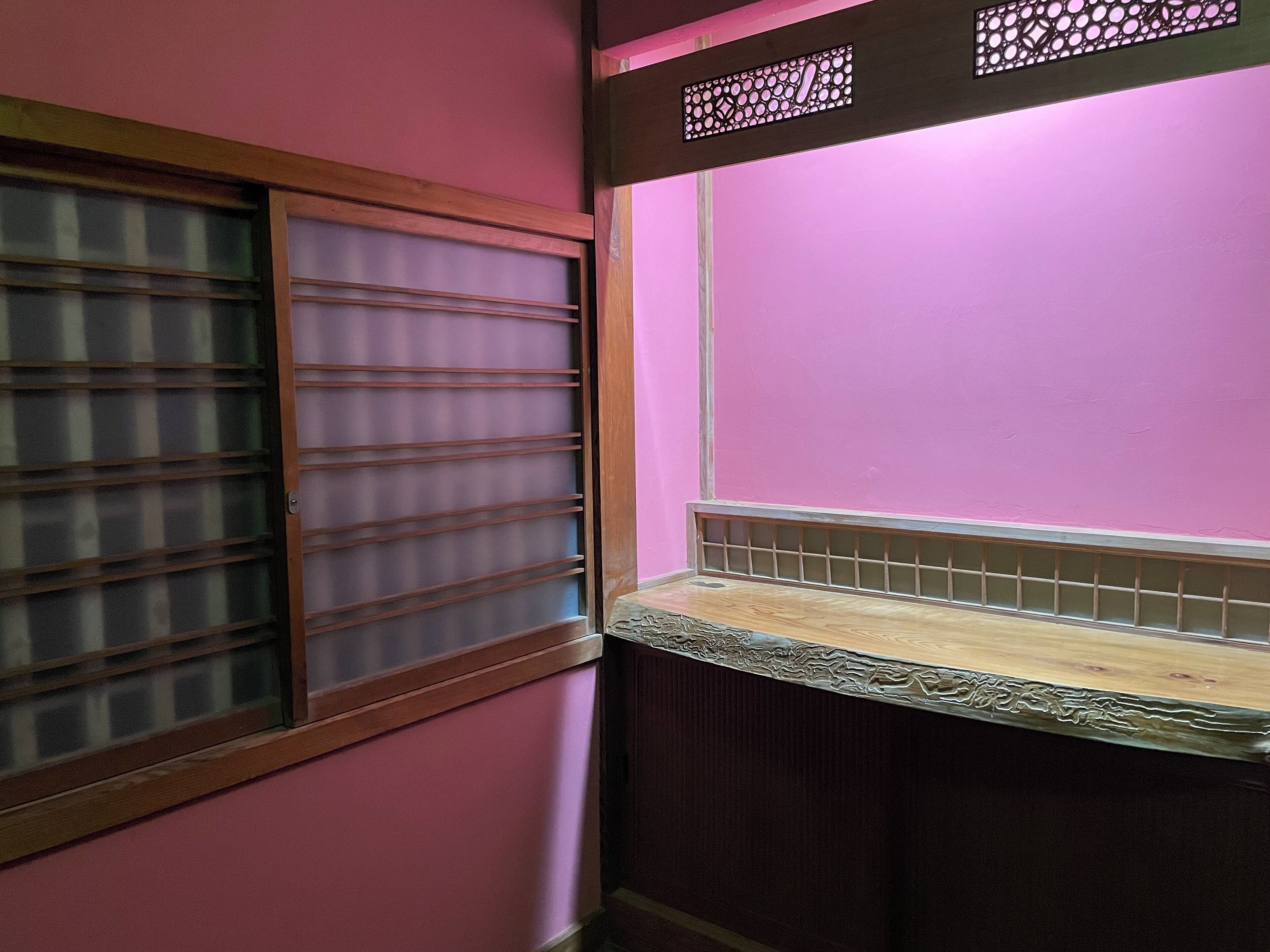 A room with a pink wall, a window with horizontal blinds, and a wooden shelf.