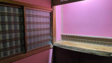 A room with a pink wall, a window with horizontal blinds, and a wooden shelf.