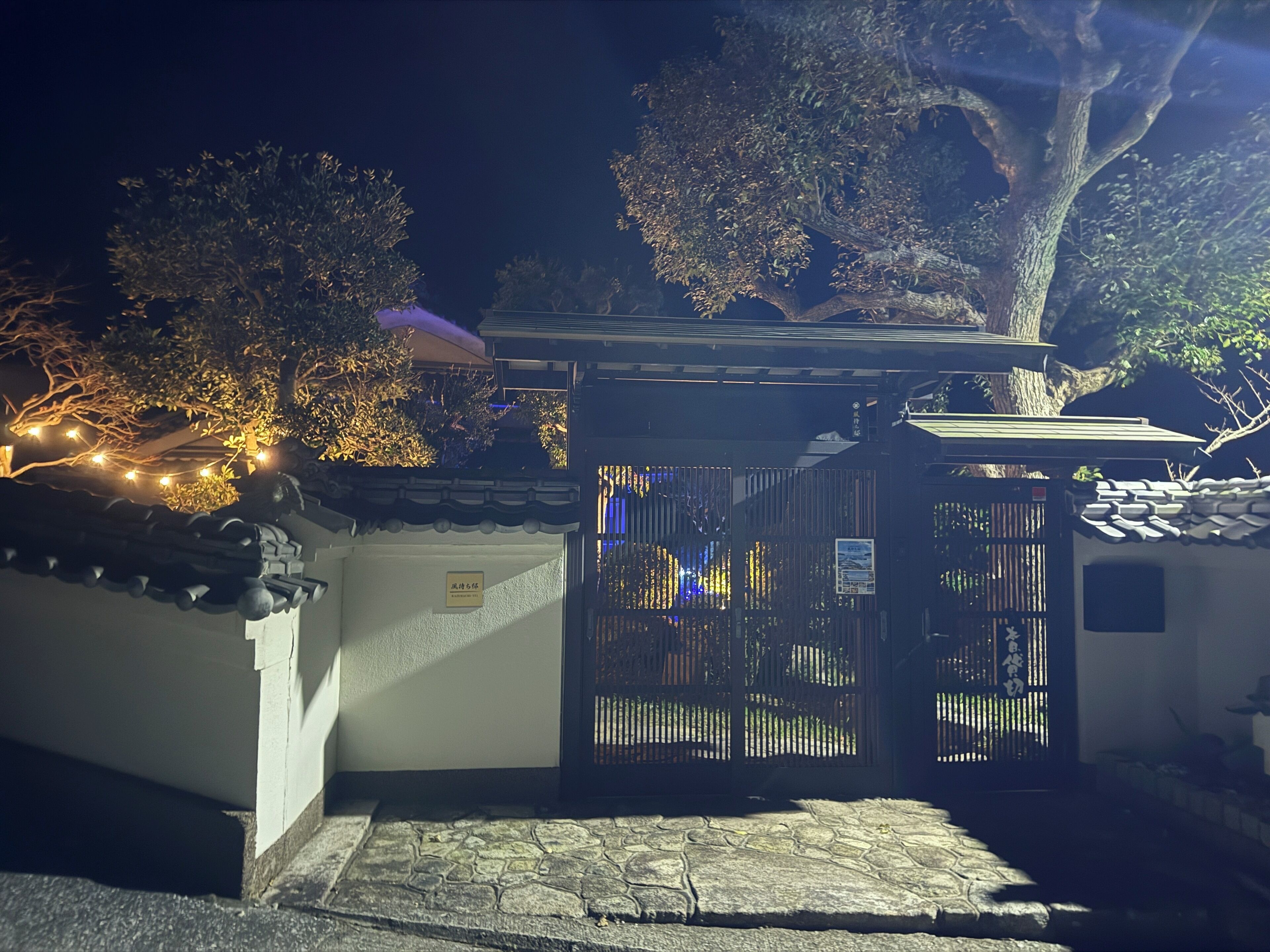 A traditional Japanese gate with a stone pathway leading to it, surrounded by trees and a building with a tiled roof.