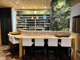 A bar area with a counter, white chairs, and shelves with various items.