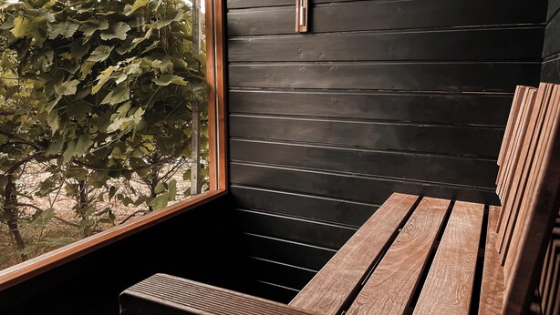 Un banc en bois doté d’une structure métallique, une fenêtre donnant sur un feuillage verdoyant et un mur en bois noir.